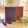 Two bars of Oatmeal & Aloe | Lavender & Goats Milk soap with lavender flowers and aloe slices on a marble surface