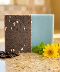 Arnica & Mint | Exfoliating Coffee & Oatmeal soap bars beside coffee beans, yellow flowers, and green mint leaves on countertop