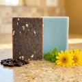 Arnica & Mint | Exfoliating Coffee & Oatmeal soap bars beside coffee beans, yellow flowers, and green mint leaves on countertop